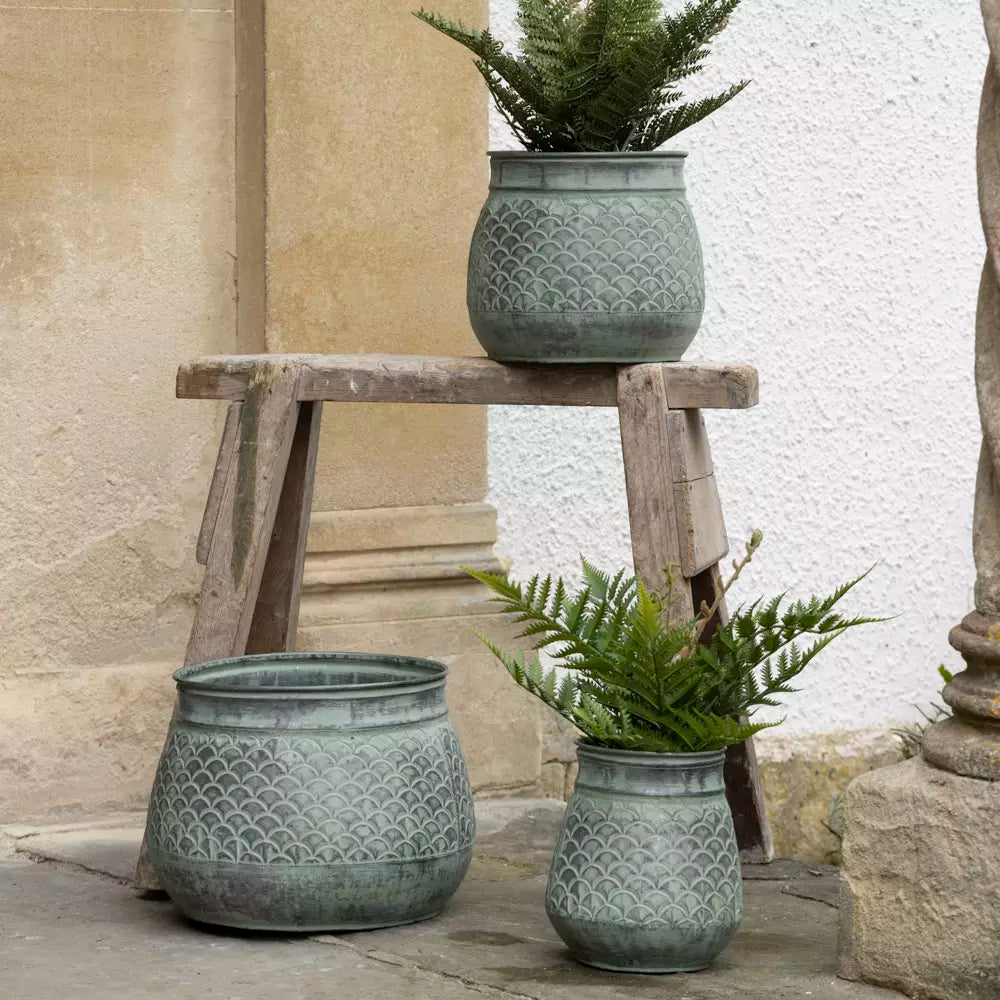 A round galvanised metal decorative plant pot in gray color, with a plant inside, placed on a stone surface.